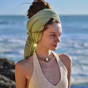 May include: A light green textured head wrap styled around the hair, paired with a cream-coloured sleeveless top featuring a V-neckline and a metallic choker necklace with a central white stone pendant, worn against a coastal background.