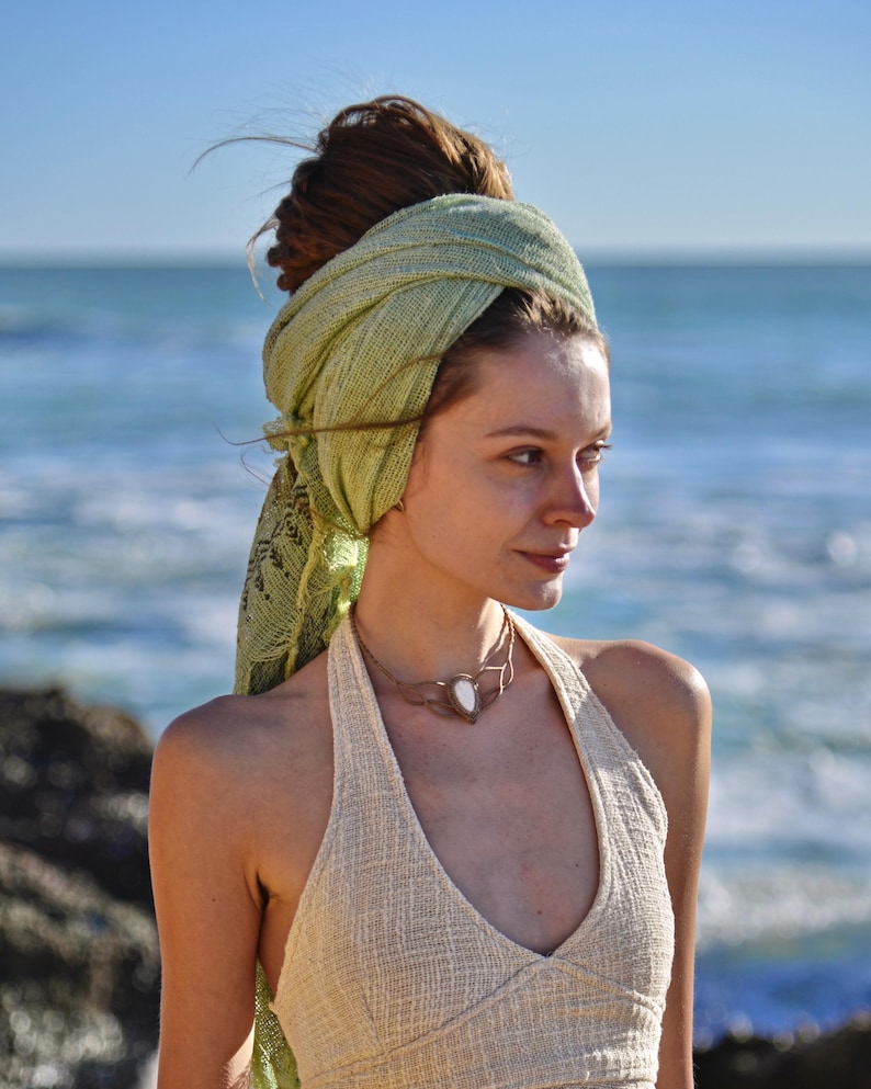 May include: A light green textured head wrap styled around the hair, paired with a cream-coloured sleeveless top featuring a V-neckline and a metallic choker necklace with a central white stone pendant, worn against a coastal background.