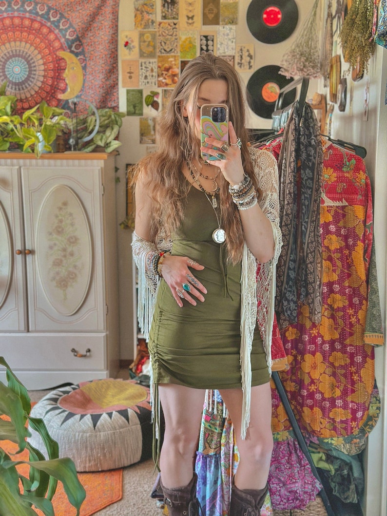 May include: A woman wearing a green, fitted, mini dress with fringe detailing. She is wearing a white crocheted cardigan and several bracelets on her wrist. She is standing in a room with a white cabinet and a patterned rug.