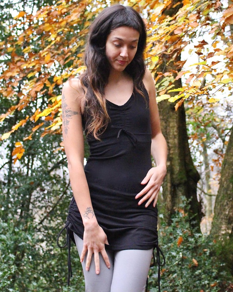 May include: A black, sleeveless, gathered top with adjustable straps and a drawstring hem. The top is worn with grey pants and is photographed in a forest setting.