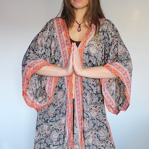 May include: A woman wearing a long, flowing black and orange patterned robe with a long-sleeved design. The robe has a floral pattern and a tie closure at the waist. The woman is standing with her hands clasped in front of her.