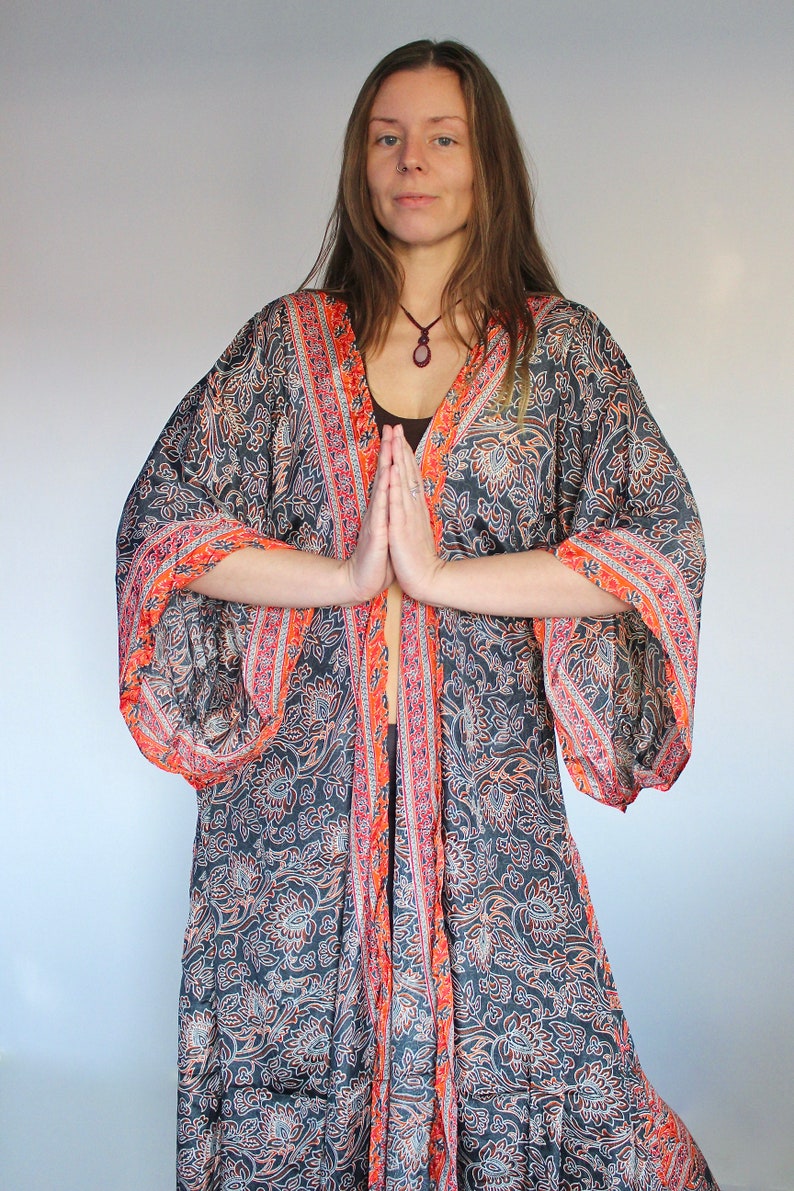 May include: A woman wearing a long, flowing black and orange patterned robe with a long-sleeved design. The robe has a floral pattern and a tie closure at the waist. The woman is standing with her hands clasped in front of her.