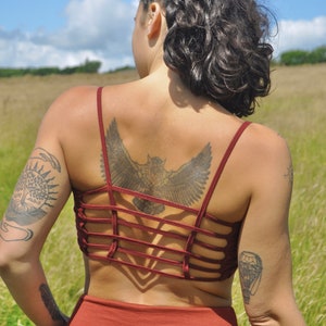 May include: A woman with a braided hairstyle wears a maroon crop top with a criss-cross back design. She has a large owl tattoo on her back and other tattoos on her arms.