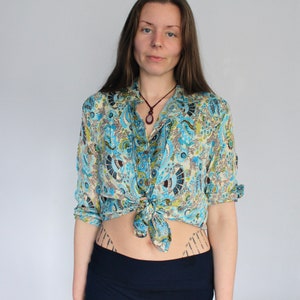 Op de afbeelding: Een blauw-wit gebloemd overhemd met lange mouwen en een knoopsluiting. Het overhemd is in de taille geknoopt en het model draagt een donkerblauwe rok.