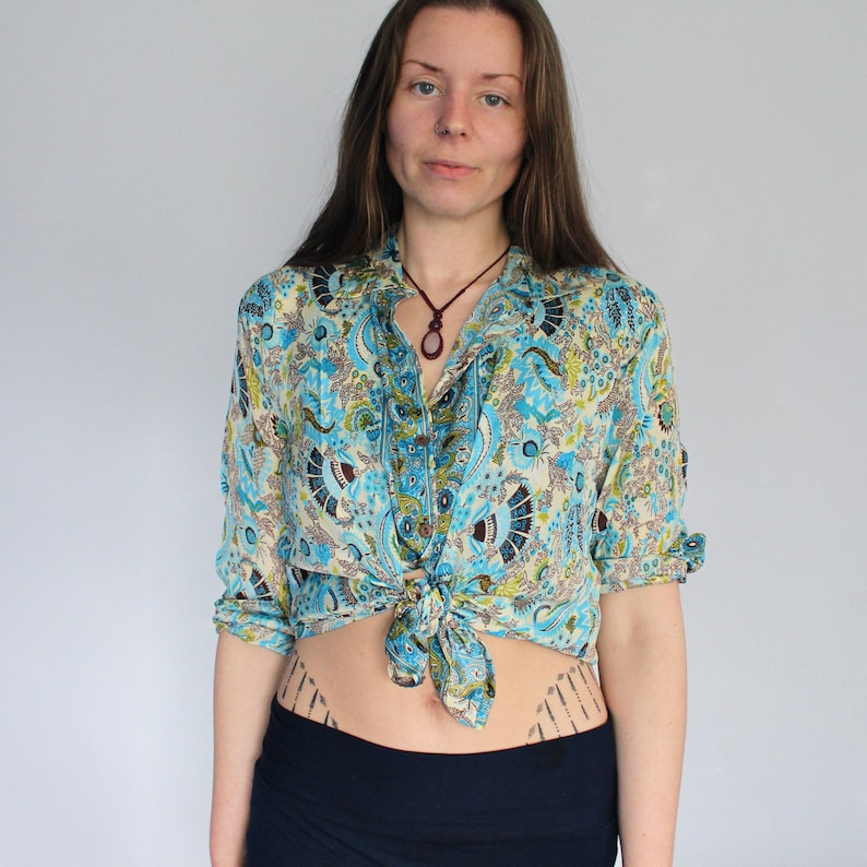 Op de afbeelding: Een blauw-wit gebloemd overhemd met lange mouwen en een knoopsluiting. Het overhemd is in de taille geknoopt en het model draagt een donkerblauwe rok.