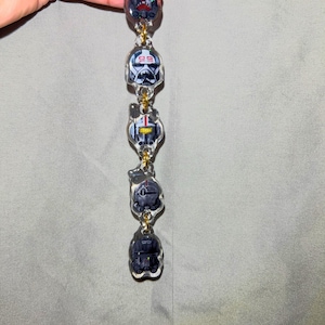 Könnte beinhalten: Ein klarer Acryl-Anhänger mit fünf detaillierten Illustrationen von Star Wars-Helmen, die durch goldfarbene Ketten verbunden sind. Die Helme sind in Schwarz-, Weiß- und Rottönen gehalten und weisen komplizierte Designs auf. Der Anhänger wird vor einem grauen Hintergrund gehalten.