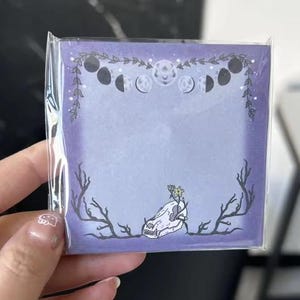 Puede incluir: Un bloc de notas cuadrado, morado, con un diseño místico. El diseño incluye fases lunares, una calavera y ramas. El bloc de notas se guarda en un envoltorio de plástico transparente.