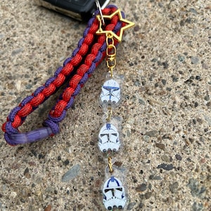 May include: A red and purple paracord keychain with a gold star charm and three clear charms featuring images of Star Wars clone trooper helmets.