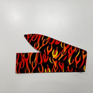May include: A black headband with a repeating flame pattern in shades of red, orange, and yellow. The fabric is folded and laid flat against a white surface. The headband is designed to be worn around the head.