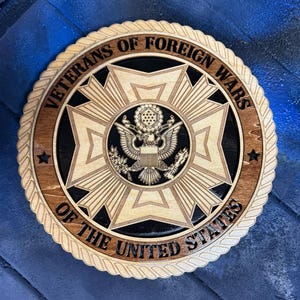 May include: A wooden plaque featuring the emblem of the Veterans of Foreign Wars. The design includes a central eagle, surrounded by a starburst pattern, and the text "Veterans of Foreign Wars of the United States". The plaque has a rope-like border.