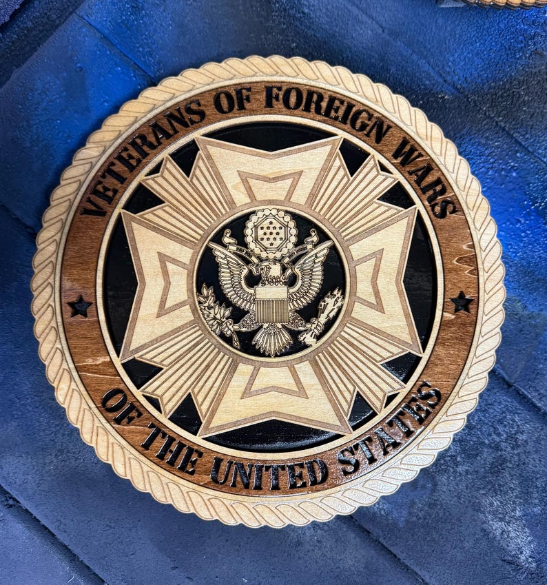 May include: A wooden plaque featuring the emblem of the Veterans of Foreign Wars. The design includes a central eagle, surrounded by a starburst pattern, and the text "Veterans of Foreign Wars of the United States". The plaque has a rope-like border.