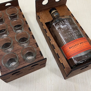 May include: Two wooden serving trays, one holding eight shot glasses and the other a bottle of Bulleit Bourbon. The trays are made of dark brown wood with cut-out designs. The bourbon bottle has an orange label.