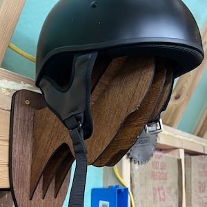 Könnte beinhalten: Ein schwarzer Motorradhelm mit einem schwarzen Riemen, der an einer Wandhalterung aus Holz hängt. Die Halterung ist handförmig mit drei Fingern.