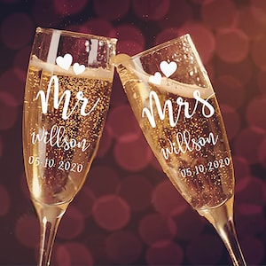 May include: Two champagne flutes with the words "Mr. Wilson 05.10.2020" and "Mrs. Wilson 05.10.2020" written on them.  The flutes are filled with champagne and have small white hearts on them.