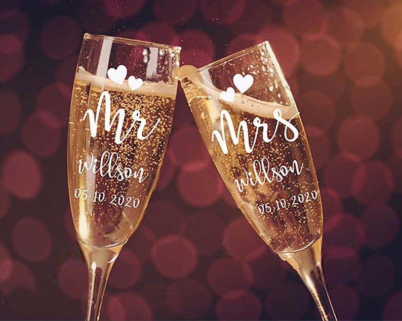 May include: Two champagne flutes with the words "Mr. Wilson 05.10.2020" and "Mrs. Wilson 05.10.2020" written on them.  The flutes are filled with champagne and have small white hearts on them.