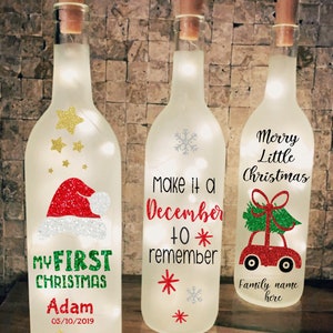 Puede incluir: Tres botellas de vidrio transparente con tapas de corcho y luces blancas en su interior. Las botellas están decoradas con purpurina y diseños navideños festivos. La primera botella dice "My First Christmas Adam 05/10/2019". La segunda botella dice "Make it a December to remember". La tercera botella dice "Merry Little Christmas Family name here".