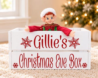 May include: A white wooden crate with red text reading Gillie's Christmas Eve Box, decorated with red snowflake icons. A small elf doll with a white hat, red sleeves, and a green collar is positioned inside the crate, looking forward.