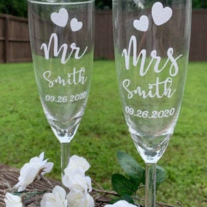May include: Two clear champagne flutes with white etched hearts and the words "Mr. Smith 09.26.2020" and "Mrs. Smith 09.26.2020".