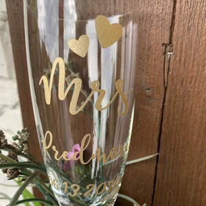 May include: A clear glass champagne flute with gold lettering that reads "Mrs. Fredmore 06.13.202" and two gold hearts.