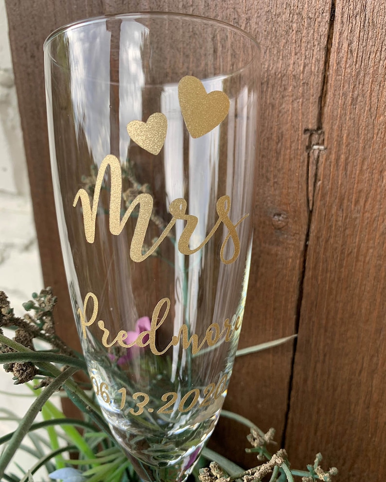 May include: A clear glass champagne flute with gold lettering that reads "Mrs. Fredmore 06.13.202" and two gold hearts.