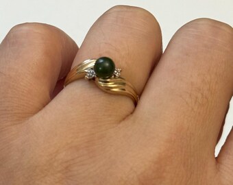 May include: A gold ring featuring a dark green gemstone and two small accent diamonds. The ring has a flowing, wave-like design on the band.