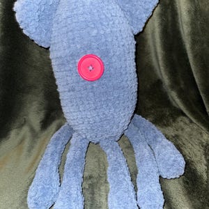 May include: A handmade blue plush toy with a button eye and ear-like appendages. The soft, textured toy has long, dangling limbs, and a bright pink button is sewn onto its body. The toy is designed to resemble a friendly creature.
