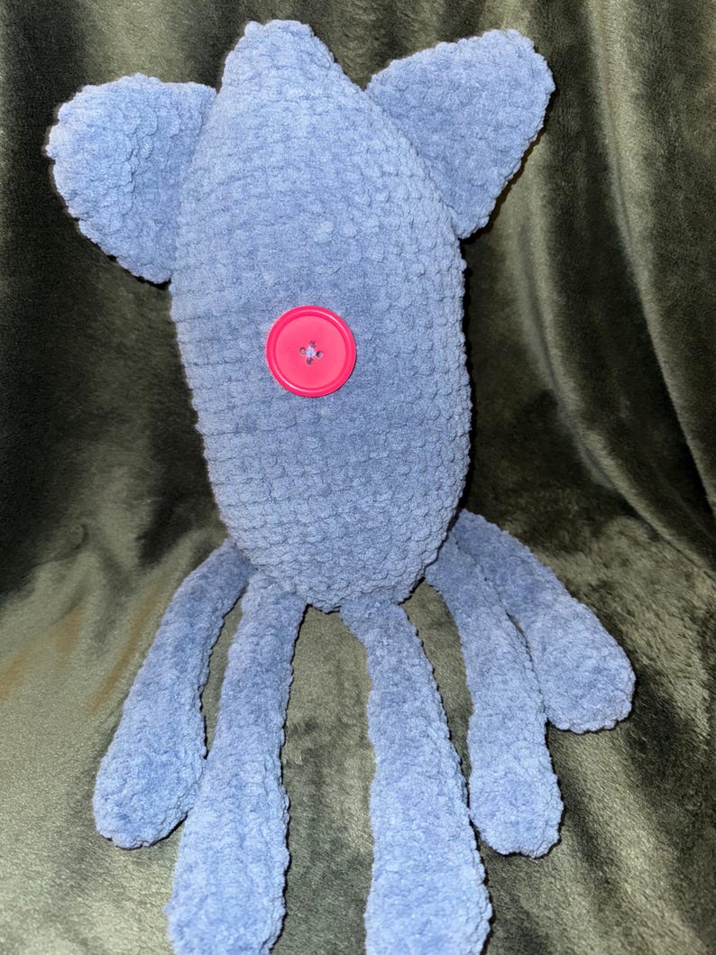 May include: A handmade blue plush toy with a button eye and ear-like appendages. The soft, textured toy has long, dangling limbs, and a bright pink button is sewn onto its body. The toy is designed to resemble a friendly creature.