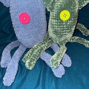 May include: Two handmade crochet monster plushies. One is blue with a pink button eye, the other is green with a yellow button eye. Both have long, tentacle-like limbs. The plushies are made of a soft, textured yarn.