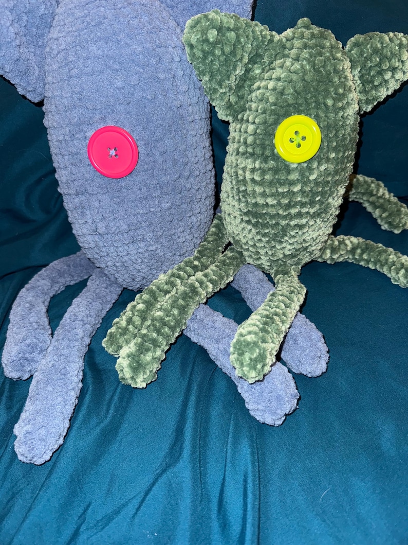 May include: Two handmade crochet monster plushies. One is blue with a pink button eye, the other is green with a yellow button eye. Both have long, tentacle-like limbs. The plushies are made of a soft, textured yarn.