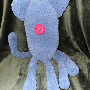 May include: A handmade, plush, blue creature with large ears, a single pink button eye, and multiple tentacle-like legs. The soft, fuzzy texture and whimsical design suggest a toy or decorative item.