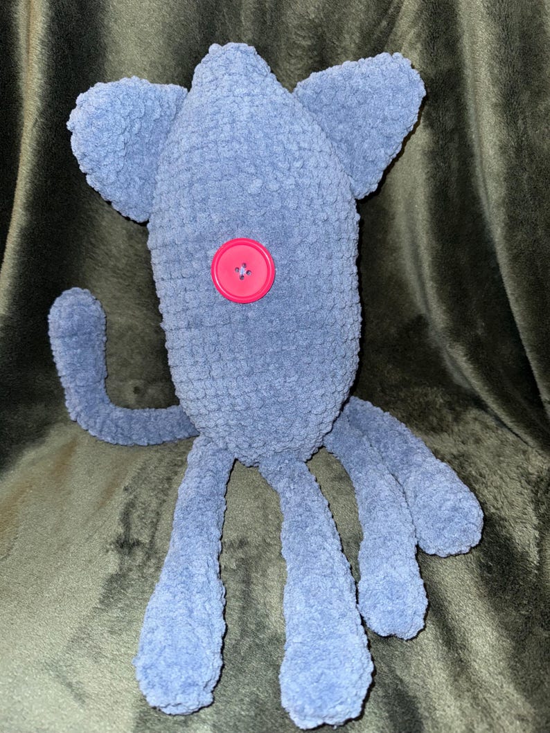 May include: A handmade, plush, blue creature with large ears, a single pink button eye, and multiple tentacle-like legs. The soft, fuzzy texture and whimsical design suggest a toy or decorative item.