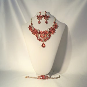 May include: A red and gold jewelry set, including a necklace, earrings, and bracelet, displayed on a white mannequin. The necklace features a floral design with a teardrop pendant. The earrings are flower-shaped, and the bracelet has a matching design.
