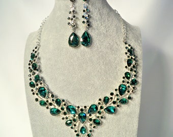 May include: A silver necklace and earrings set with emerald green teardrop-shaped crystals. The necklace features a cascading design with smaller crystals surrounding the larger teardrops.