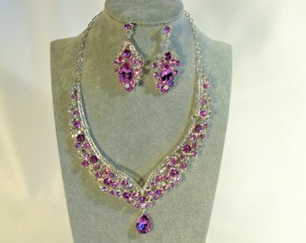 May include: A silver necklace and earrings set with a teardrop pendant and purple rhinestones.