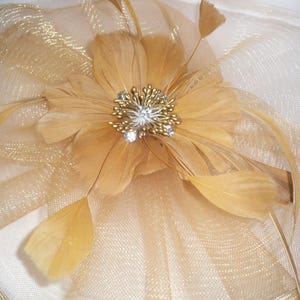 May include: A gold-colored headpiece with a central flower made of feathers and a sparkling jewel center. The headpiece has a layered design with gold mesh and feather accents, suitable for special occasions.