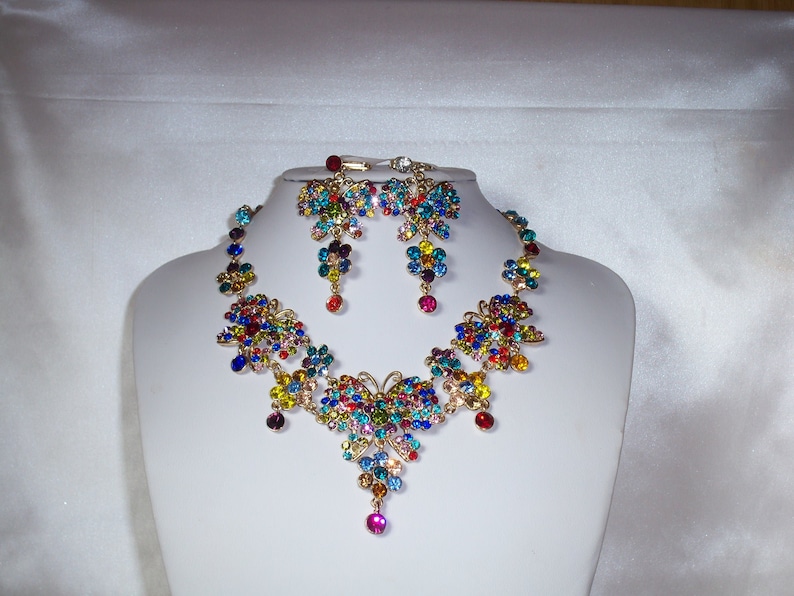 Puede incluir: Un conjunto de collar y pendientes de mariposa de colores con detalles dorados. La joyer&iacute;a presenta una variedad de piedras de cristal multicolores.