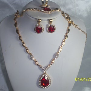 May include: A gold necklace and earrings set with a large red teardrop-shaped stone surrounded by smaller clear stones. The necklace has a delicate chain with a twisted design.