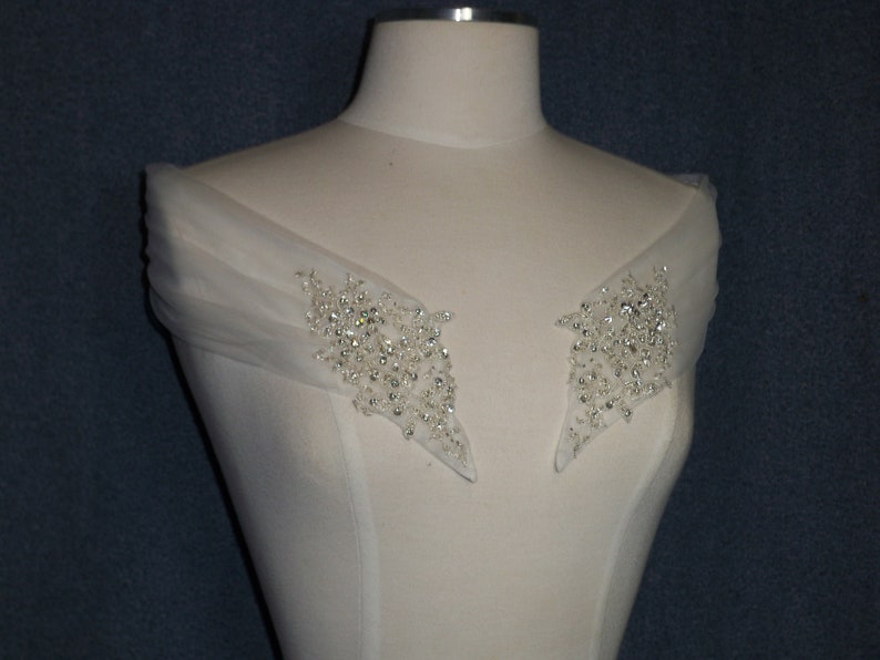 May include: A white off-the-shoulder wedding dress bodice with a sheer overlay and two beaded embellishments on the shoulders.
