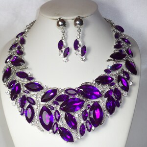 May include: A purple crystal necklace and earrings set. The necklace features a large, statement design with multiple marquise-shaped crystals. The earrings are dangle style with smaller marquise-shaped crystals.