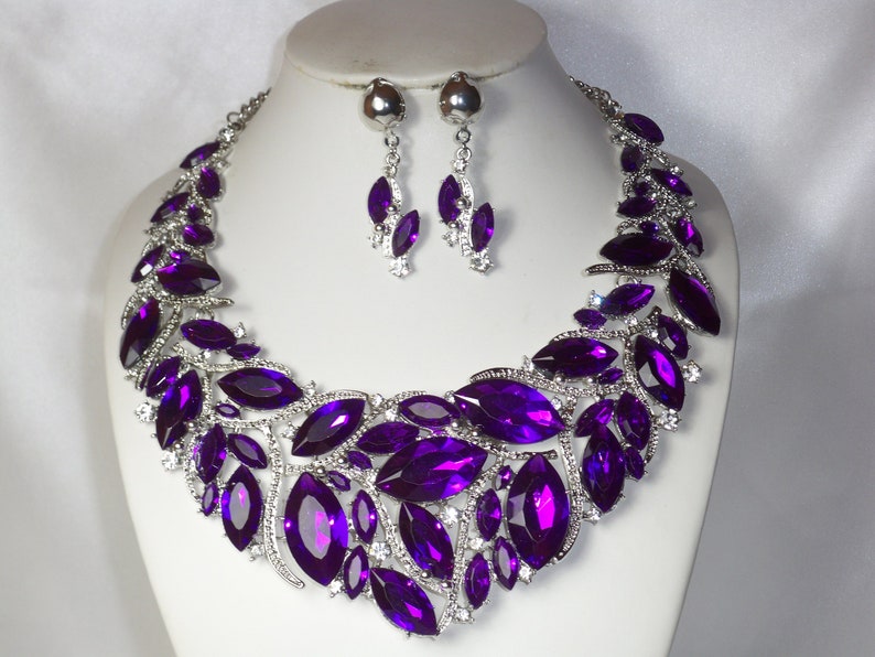 May include: A purple crystal necklace and earrings set. The necklace features a large, statement design with multiple marquise-shaped crystals. The earrings are dangle style with smaller marquise-shaped crystals.