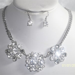 May include: A silver necklace with three flower-shaped pendants made of clear rhinestones. The necklace is paired with matching earrings with a single clear rhinestone.