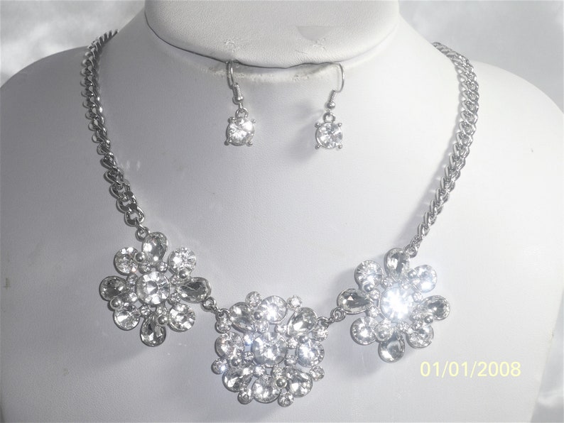 May include: A silver necklace with three flower-shaped pendants made of clear rhinestones. The necklace is paired with matching earrings with a single clear rhinestone.
