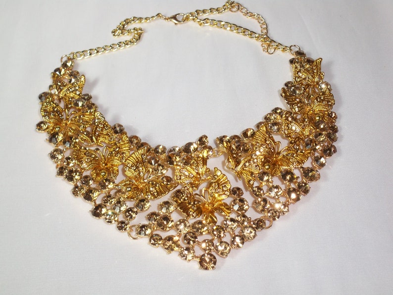 Puede incluir: Un collar dorado con dise&ntilde;os florales y de mariposas intrincados. El collar presenta numerosas piedras de estr&aacute;s redondas y transparentes y una delicada cadena para ajustar la longitud. El dise&ntilde;o es sim&eacute;trico y adecuado para ocasiones especiales.
