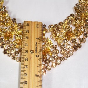 Puede incluir: Un collar dorado con dise&ntilde;os de mariposas y flores, adornado con pedrer&iacute;a transparente y &aacute;mbar. El collar mide aproximadamente 25 cm de largo, como indica la regla. Una pieza de joyer&iacute;a llamativa.