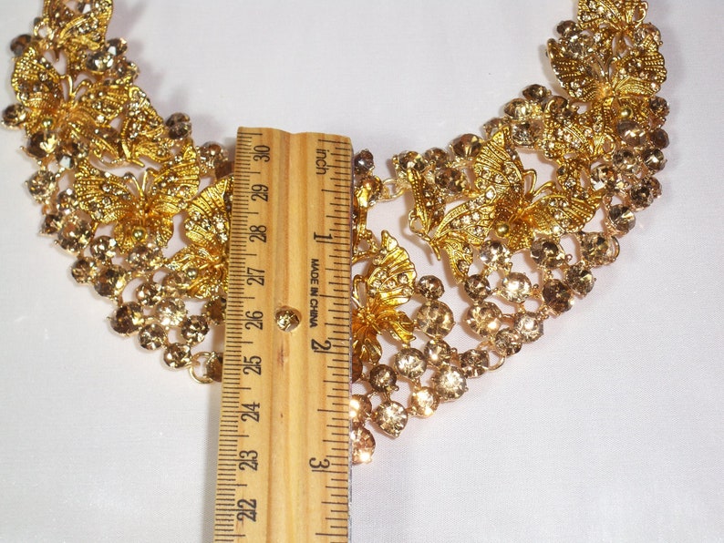 Puede incluir: Un collar dorado con dise&ntilde;os de mariposas y flores, adornado con pedrer&iacute;a transparente y &aacute;mbar. El collar mide aproximadamente 25 cm de largo, como indica la regla. Una pieza de joyer&iacute;a llamativa.