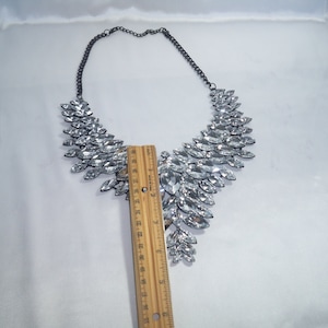Puede incluir: Un collar llamativo con numerosos cristales transparentes de talla marquesa dispuestos en un patr&oacute;n de abanico sobre una cadena de metal oscuro. Una regla de madera colocada en el centro muestra que la secci&oacute;n de cristales mide 10 cm de largo.
