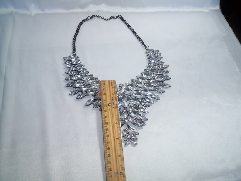Puede incluir: Un collar llamativo con numerosos cristales transparentes de talla marquesa dispuestos en un patr&oacute;n de abanico sobre una cadena de metal oscuro. Una regla de madera colocada en el centro muestra que la secci&oacute;n de cristales mide 10 cm de largo.