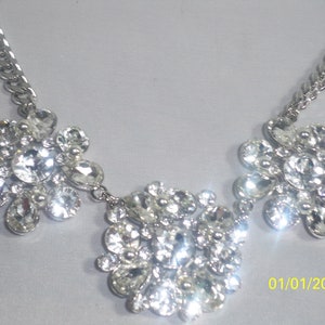 May include: A silver chain necklace with three large, clear rhinestone clusters. The clusters are arranged in a symmetrical pattern, with the center cluster being the largest.
