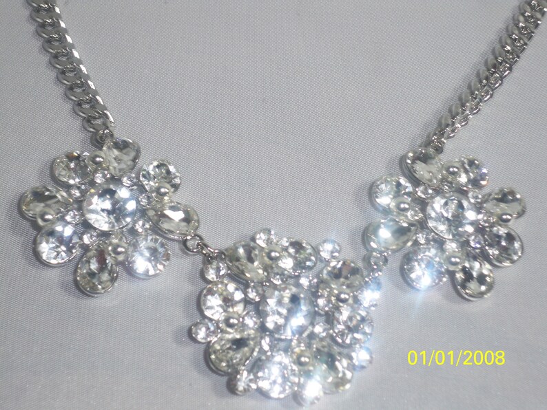 May include: A silver chain necklace with three large, clear rhinestone clusters. The clusters are arranged in a symmetrical pattern, with the center cluster being the largest.