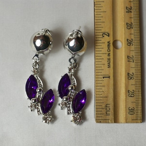 May include: A pair of silver dangle earrings with purple and clear rhinestones. The earrings are approximately 1 inch long.